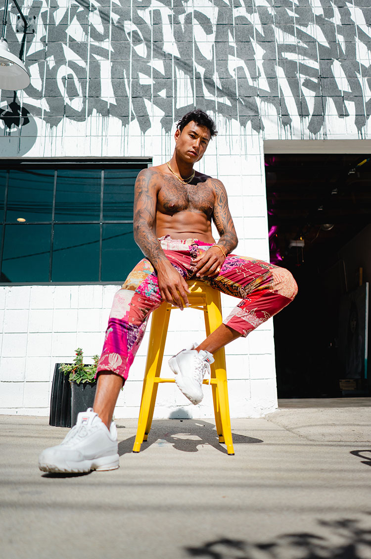 Topless man sits outside on yellow stool in front of white brick wall