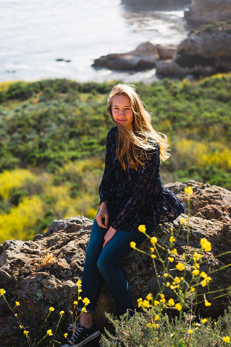 Woman sits on rock in field of yellow flowers by ocean